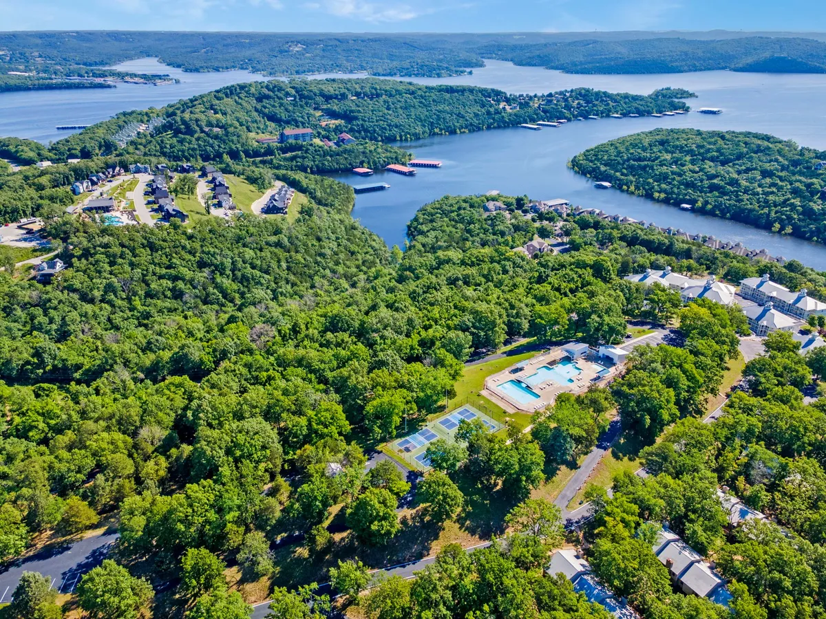 A wide view of the resort grounds featuring a swimming pool area, tennis courts (which can easily represent pickleball as well), residential buildings, and lush greenery, all set against the backdrop of a scenic lake. This comprehensive scene highlights the abundance of outdoor recreational amenities available on-site.