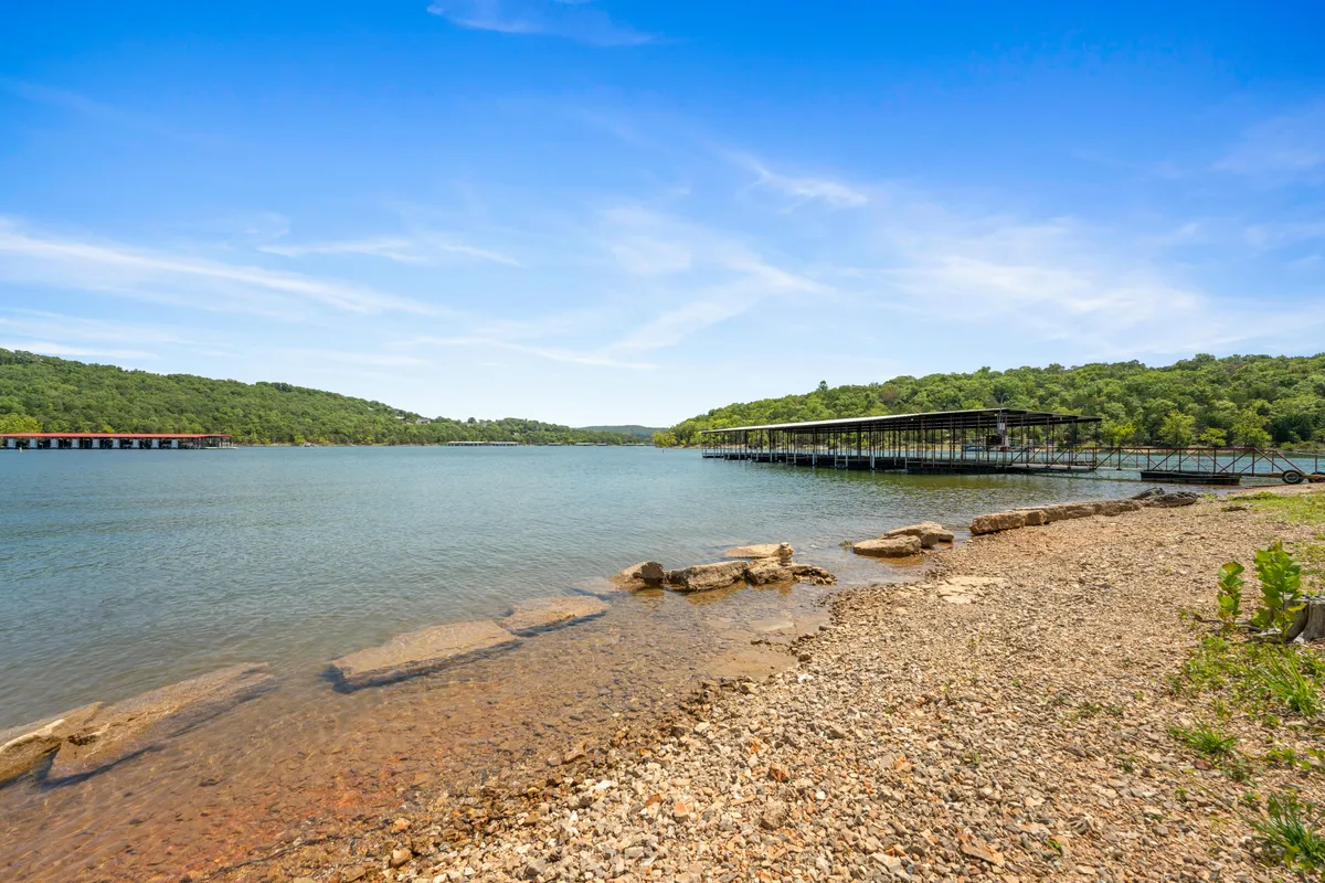 A tranquil view of Table Rock Lake in the foreground, with calm waters, a dock extending into the lake, a rocky shoreline, and distant hills on the horizon. The scene captures the serene lake atmosphere just steps from the condo and hints at activities like boating, fishing, and enjoying the surrounding natural beauty.