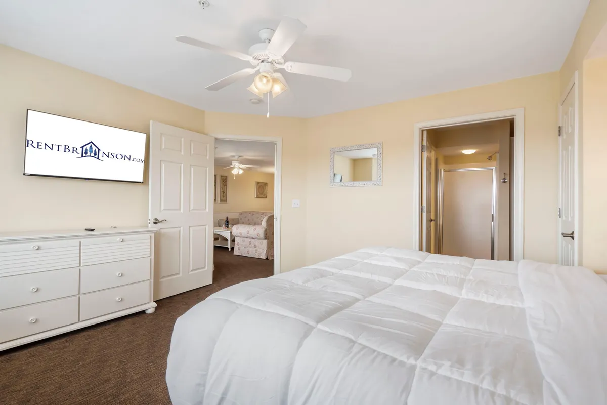 A serene bedroom scene with a neatly made bed adorned in crisp white linens, a wall-mounted TV, a dresser with a mirror, and a doorway leading to another space. The room feels quiet and inviting, with plenty of space for personal comfort and relaxation.