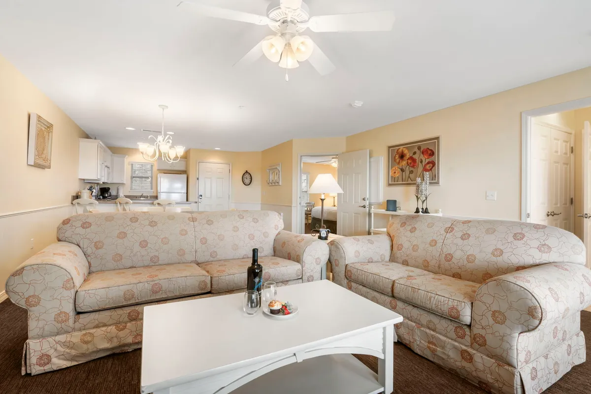 A bright, welcoming living area with plush sofas, a central coffee table, a chandelier overhead, and decorative wall art. Sunlight pours in through large windows, creating a warm and inviting atmosphere ideal for both people and pets.