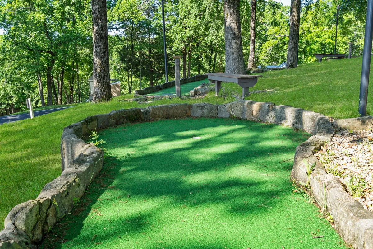 A vibrant mini-golf course nestled among mature trees, with benches for relaxing and socializing. The space exudes a playful and communal vibe perfect for family fun and friendly competition.