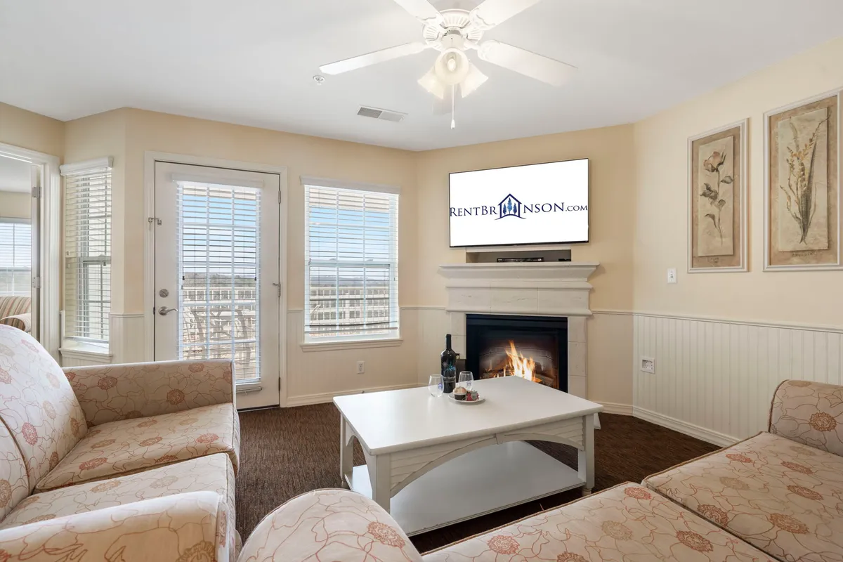 A cozy, open living area featuring a plush sofa, coffee table, TV, and an electric fireplace as the central focal point. The warm tones, soft lighting, and spacious layout evoke a sense of comfort and togetherness, perfect for relaxing or socializing.