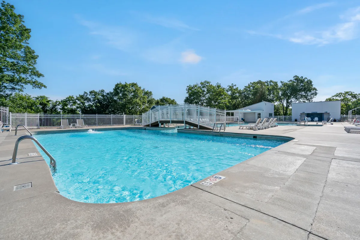 A spacious, sunlit outdoor swimming pool with a decorative bridge and plenty of lounge chairs, surrounded by lush greenery. The atmosphere is inviting, with space for socializing, swimming, and relaxing under the open sky.