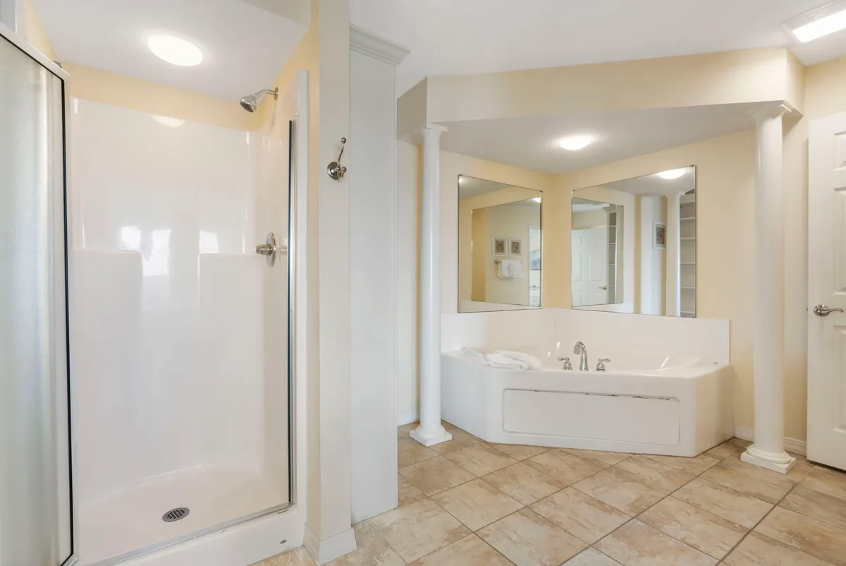 A tranquil bathroom retreat featuring a large soaker tub set against elegant columns, a separate glass-enclosed shower, and a mirrored wall that enhances the sense of space and light. The room feels spa-like and serene, ideal for unwinding after a day by the lake.