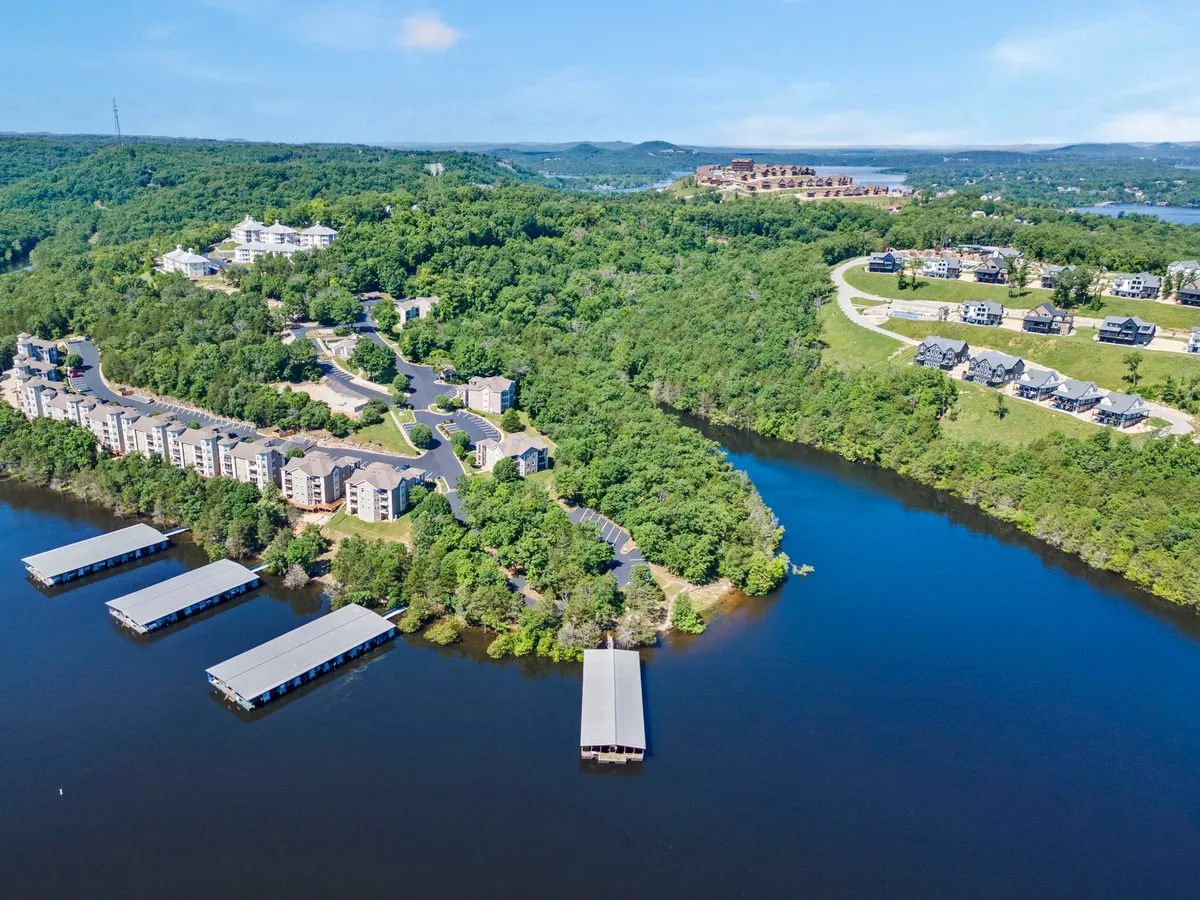 A sweeping exterior shot revealing waterfront properties nestled along the lake, with private docks, lush greenery, and gently rolling hillside developments. The scene highlights both the natural beauty and resort-style amenities available to guests.