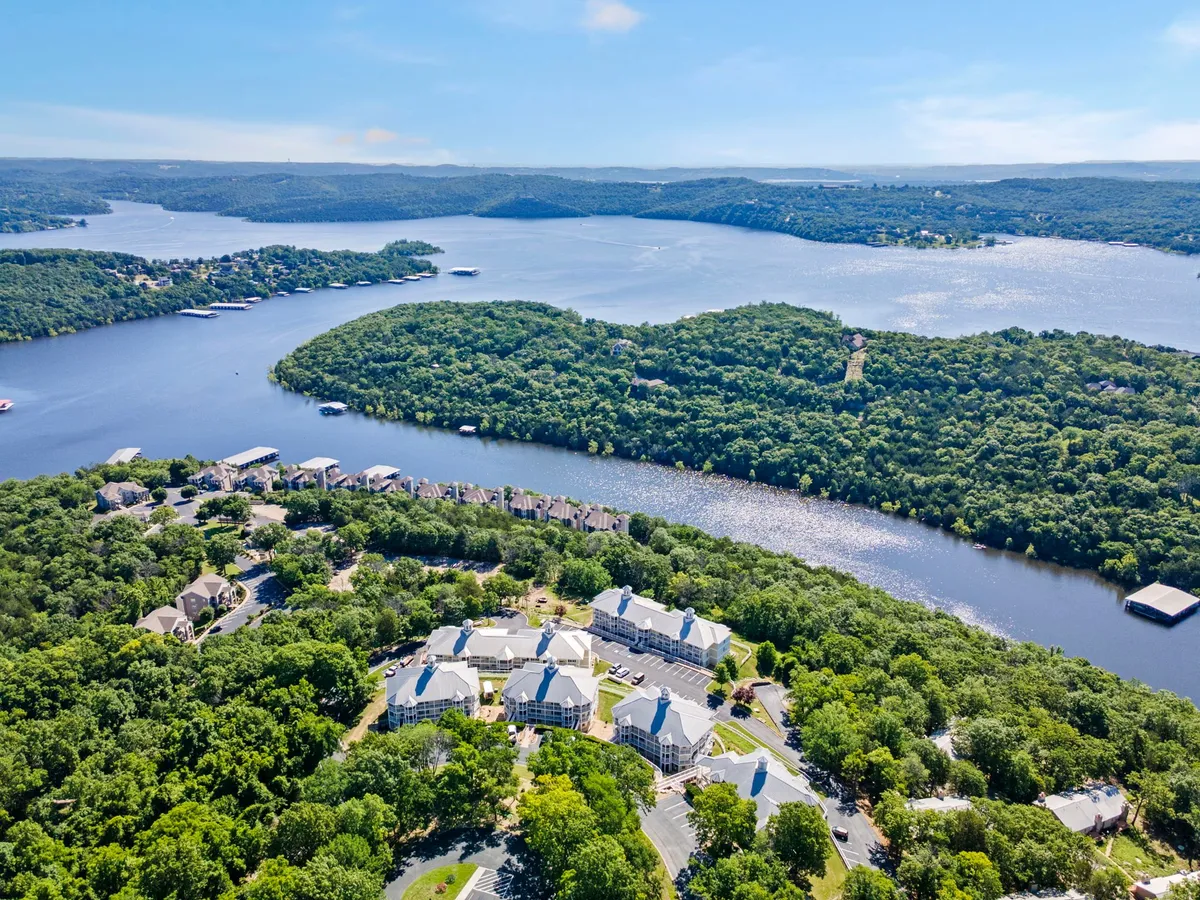 The image showcases a tranquil lakefront setting at sunset, with shimmering water, lush wooded surroundings, small islands, and glimpses of residential condo buildings nestled amid the greenery. The peaceful scene invites thoughts of boating, fishing, and enjoying outdoor gatherings by the water, with the soft glow of the sunset reflecting on the lake.